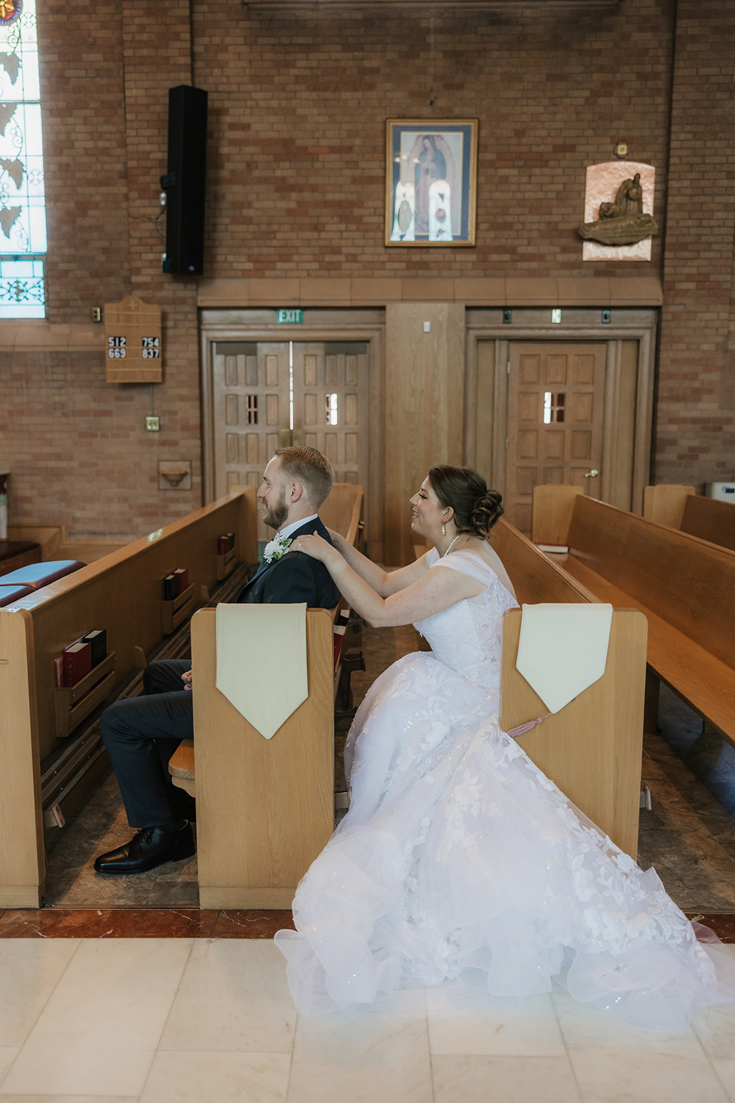 A bride and groom sit together in a church pew, facing the altar; the bride’s dress train spreads across the floor.