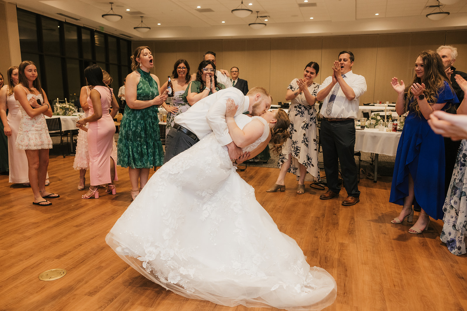 A bride and groom share a dip dance move in the center of a reception hall, surrounded by clapping and smiling guests.