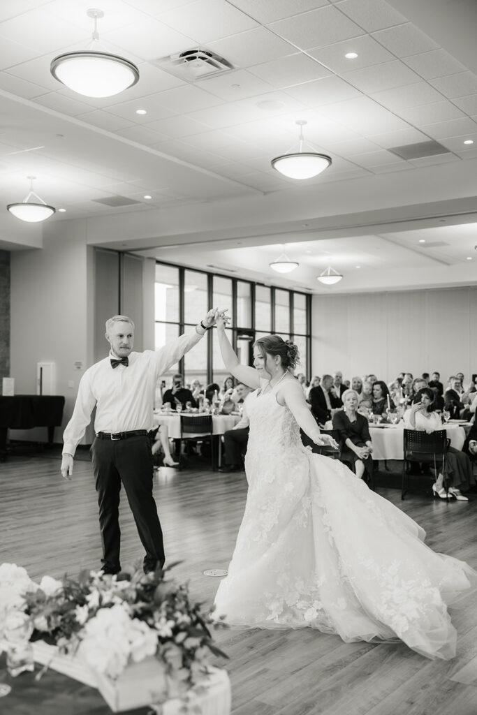 A bride and groom enter a reception hall hand in hand; the bride holds a bouquet up as guests watch and applaud.