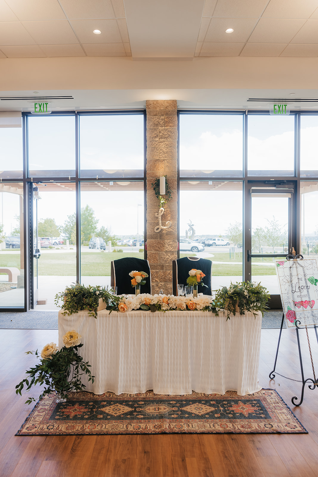 A decorated table with flowers and greenery is set in front of two chairs, positioned by large windows and glass doors in a bright indoor space.