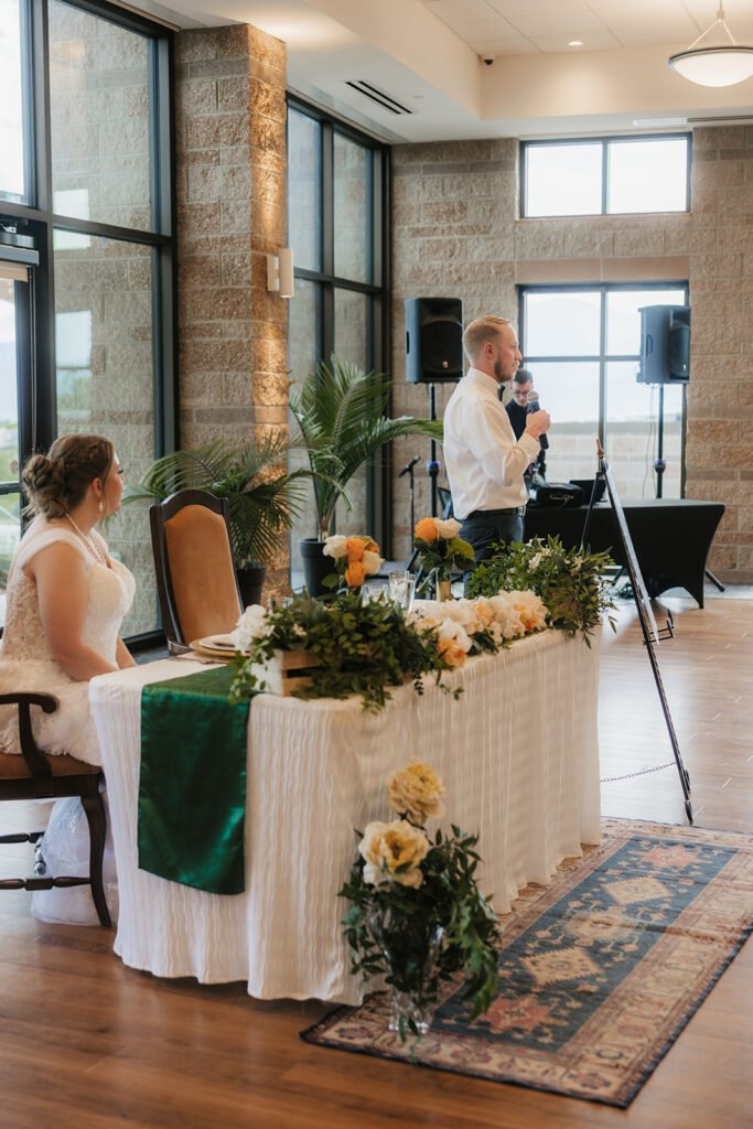 A bride and groom enter a reception hall hand in hand; the bride holds a bouquet up as guests watch and applaud.