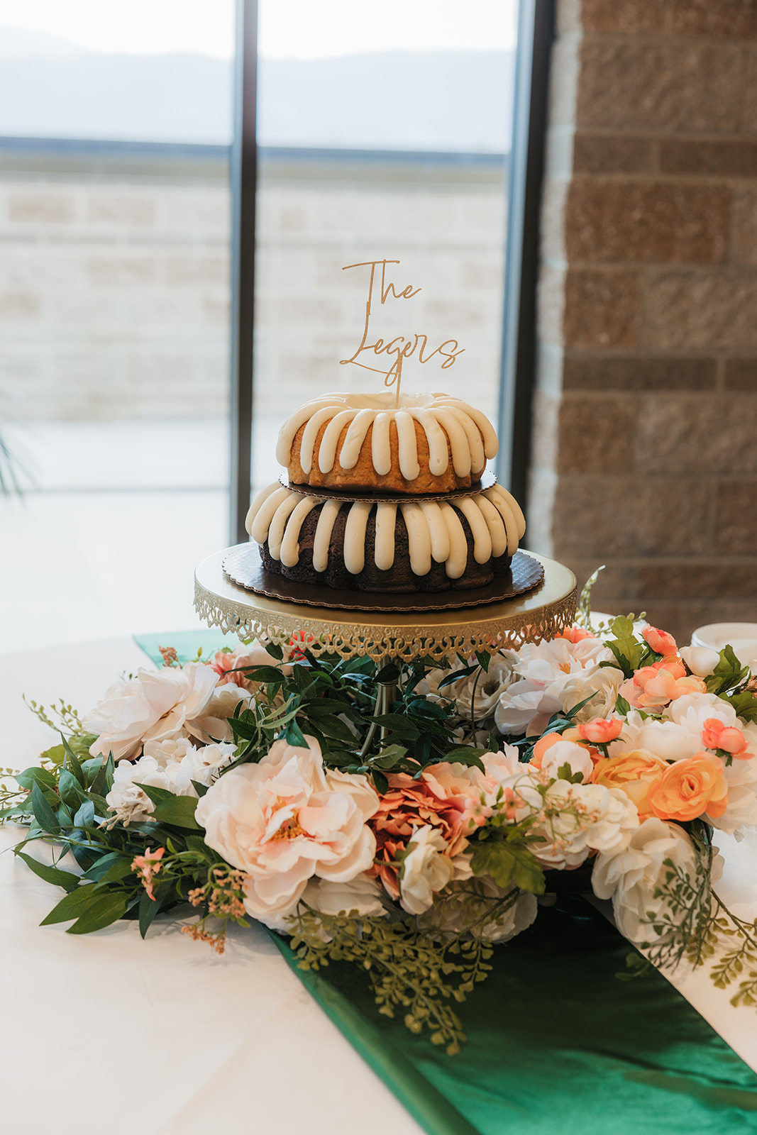 A two-tiered bundt cake with white icing sits on a cake stand, surrounded by flower arrangements and greenery on a decorated table.