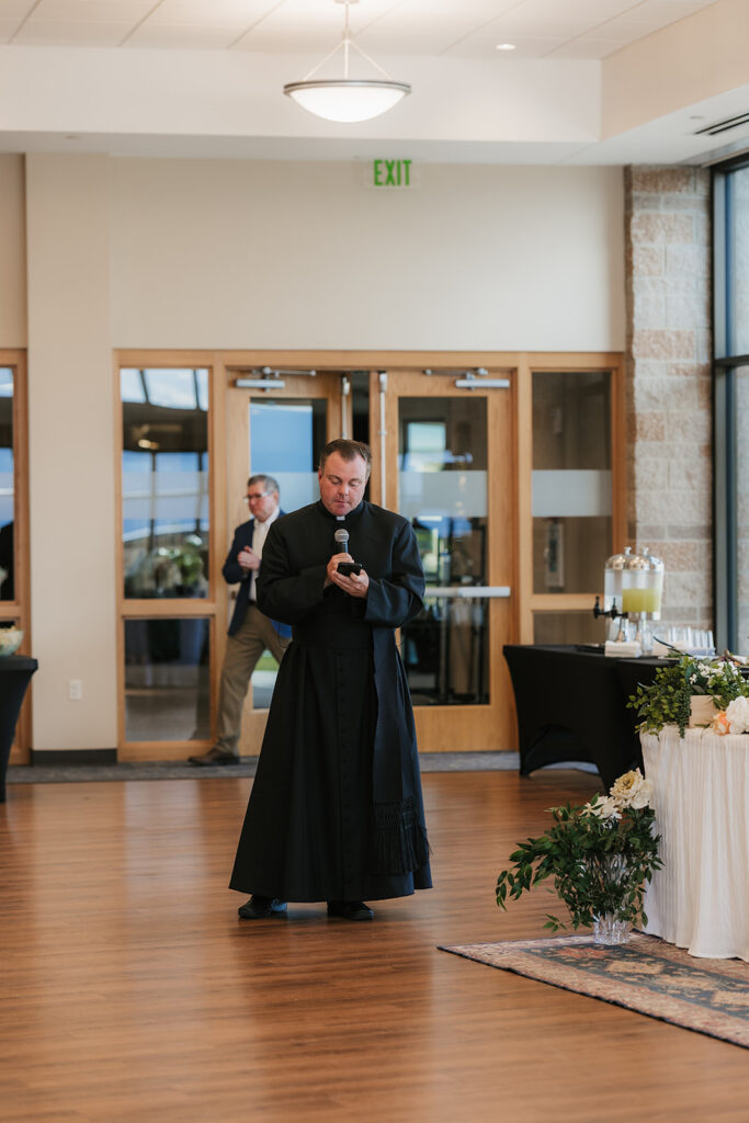 A bride and groom enter a reception hall hand in hand; the bride holds a bouquet up as guests watch and applaud.
