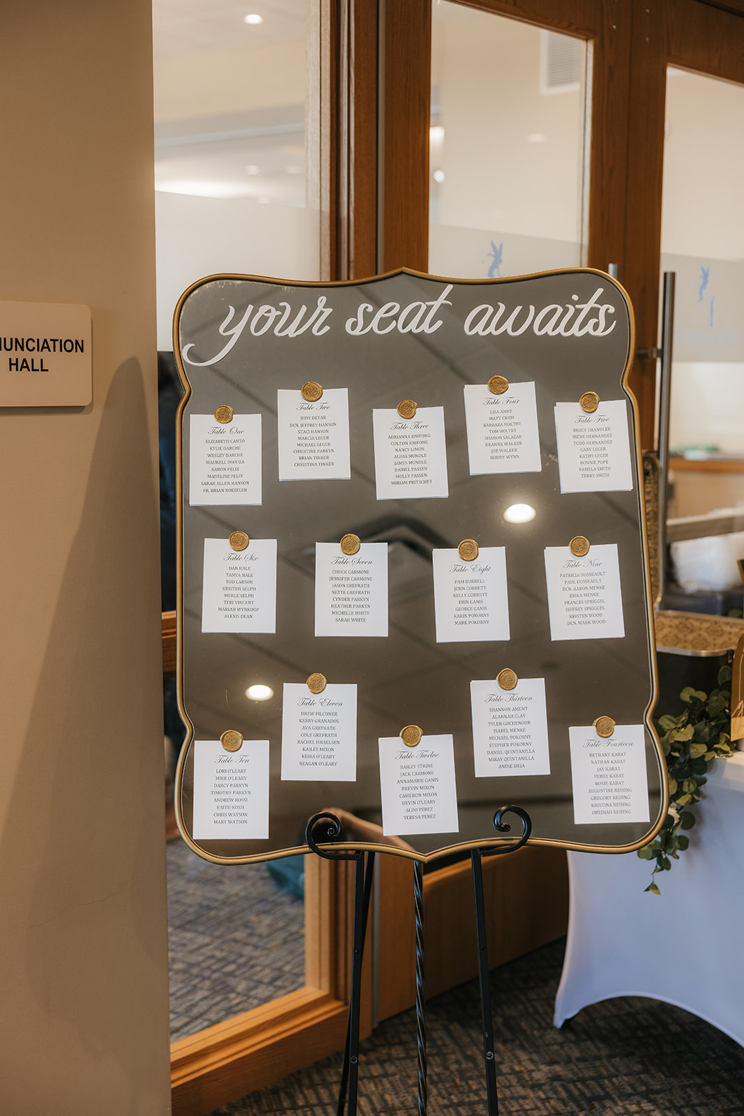 A seating chart board with the heading "your seat awaits," displaying lists of guests' names and table assignments, placed near a doorway labeled "Educator Hall.