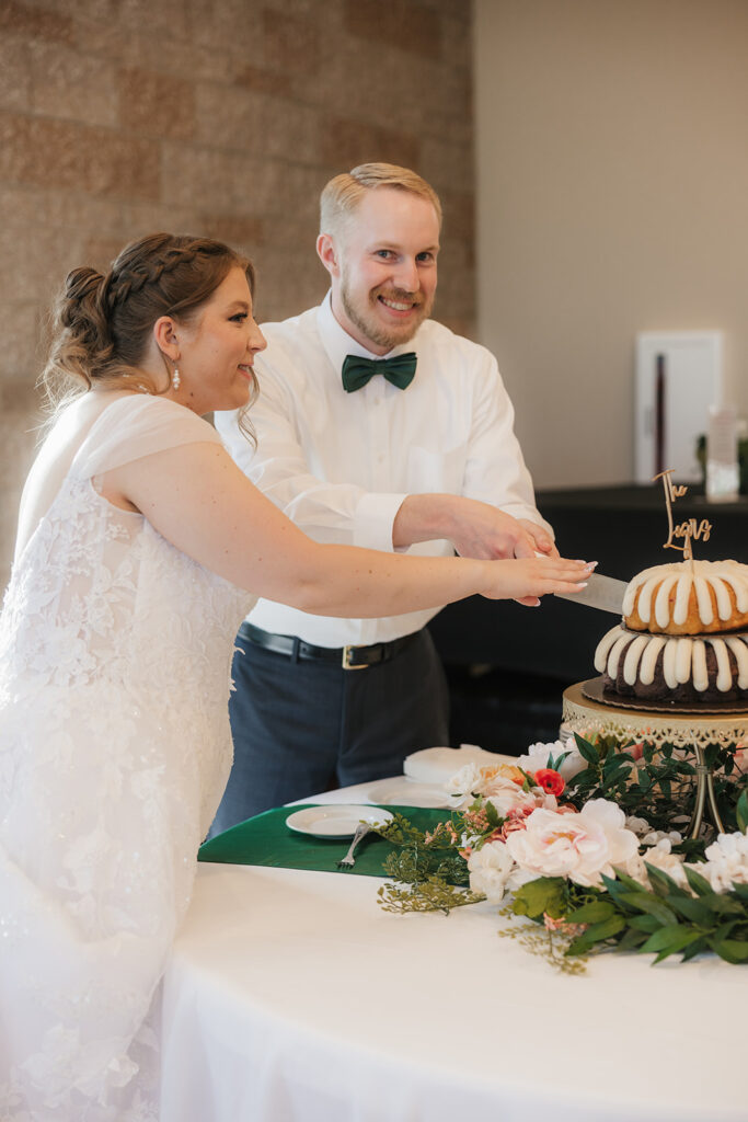 A bride and groom feed each other cake at their wedding reception, standing beside a decorated cake table with flowers and stacked bundt cakes.
