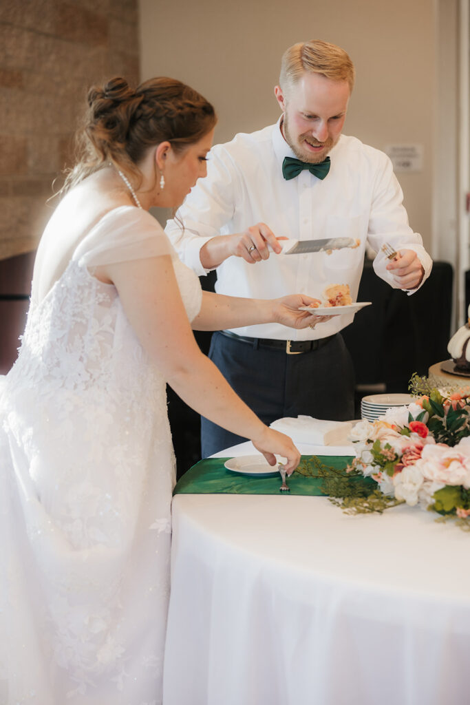 A bride and groom feed each other cake at their wedding reception, standing beside a decorated cake table with flowers and stacked bundt cakes.