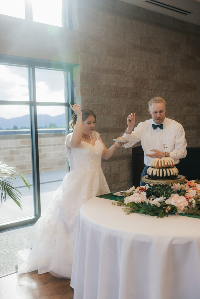 A bride and groom feed each other cake at their wedding reception, standing beside a decorated cake table with flowers and stacked bundt cakes.