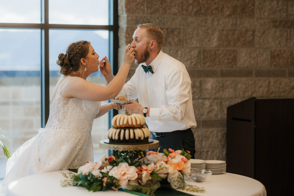 A bride and groom feed each other cake at their wedding reception, standing beside a decorated cake table with flowers and stacked bundt cakes.