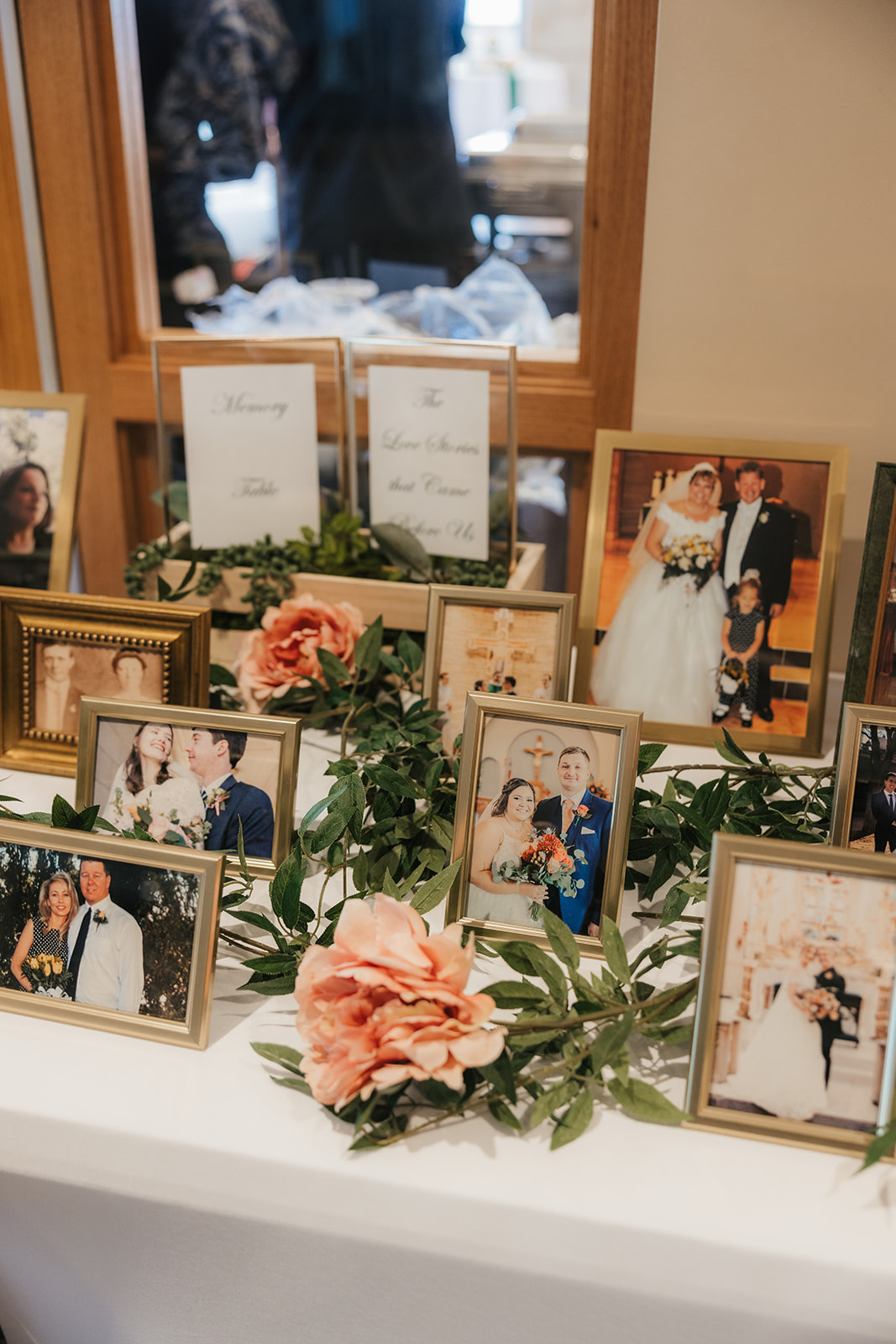 A table displays framed family photos, greenery, and pink flowers, with a window and people blurred in the background.