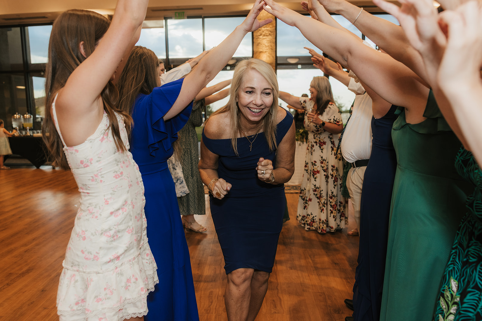 A woman in a blue dress smiles as she walks through an archway formed by people raising their arms at an indoor event.