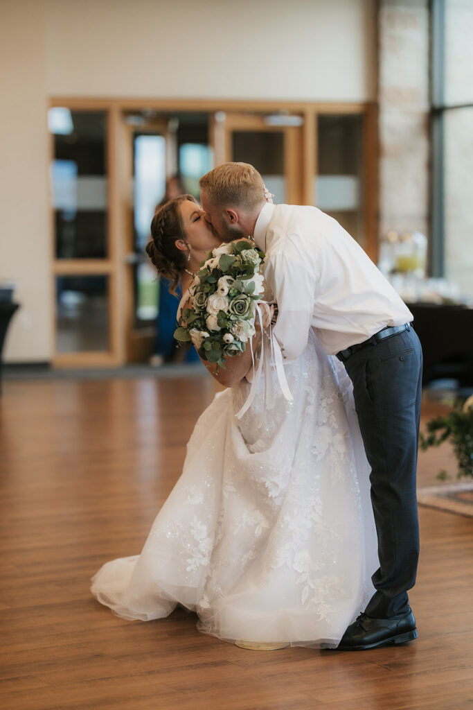 A bride and groom enter a reception hall hand in hand; the bride holds a bouquet up as guests watch and applaud.