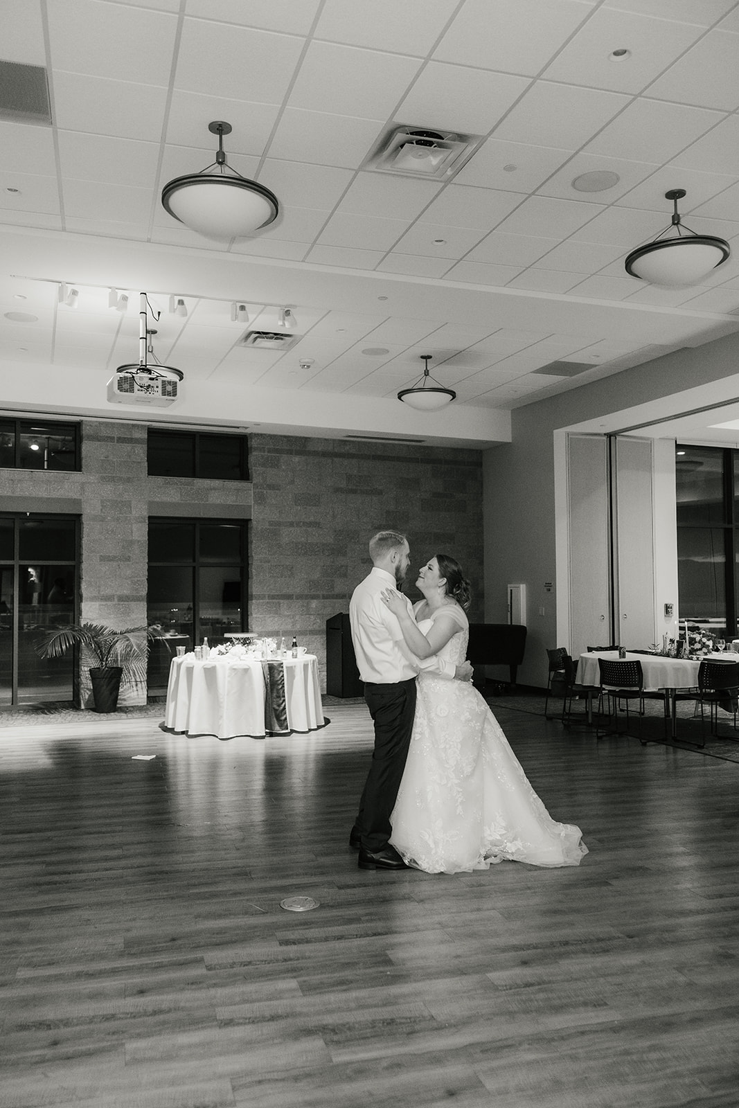 A bride and groom share a dance in the center of a large, decorated reception hall with tables and chairs arranged around them.