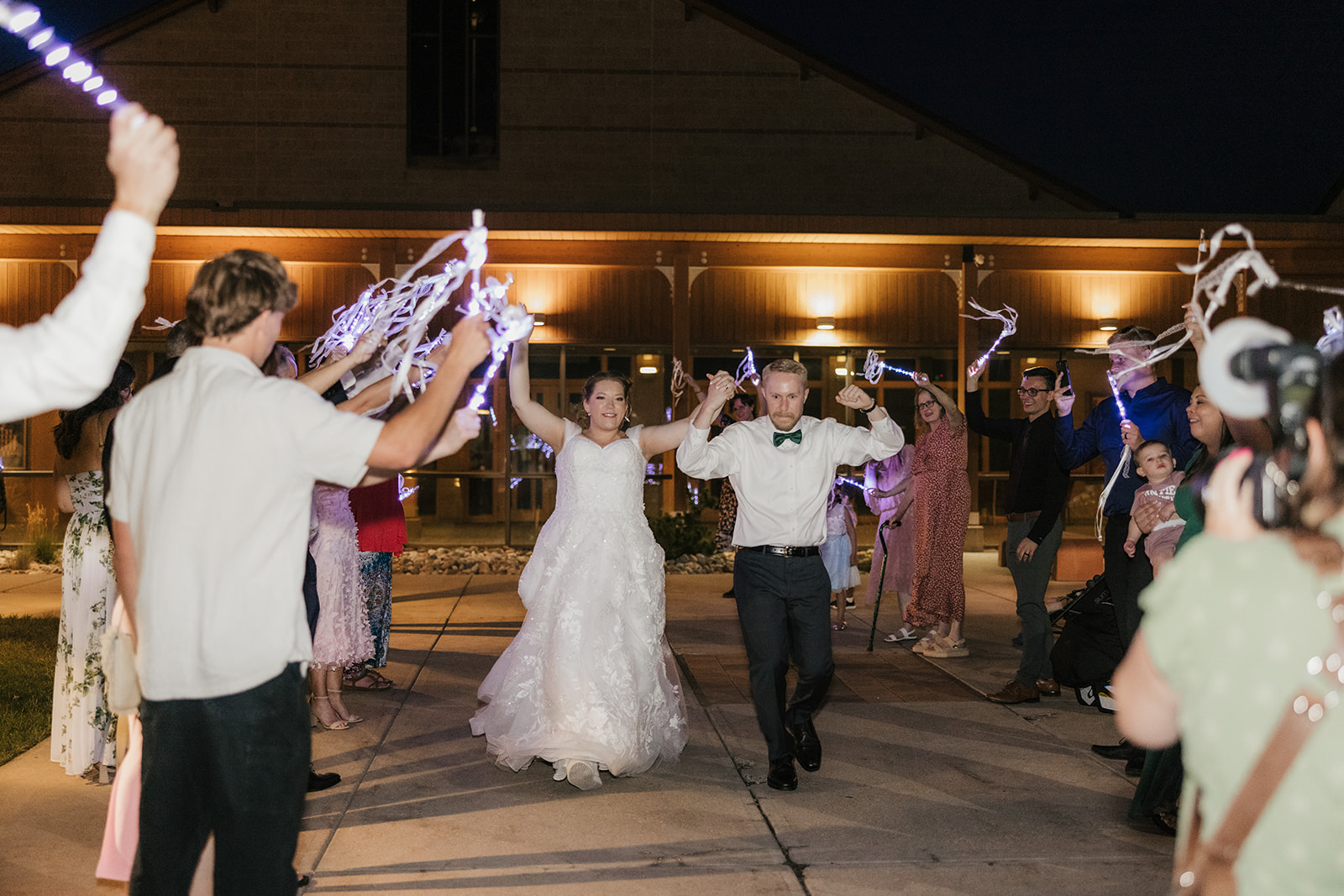 A bride and groom walk outside at night as guests hold up lights and ribbons, celebrating their wedding.