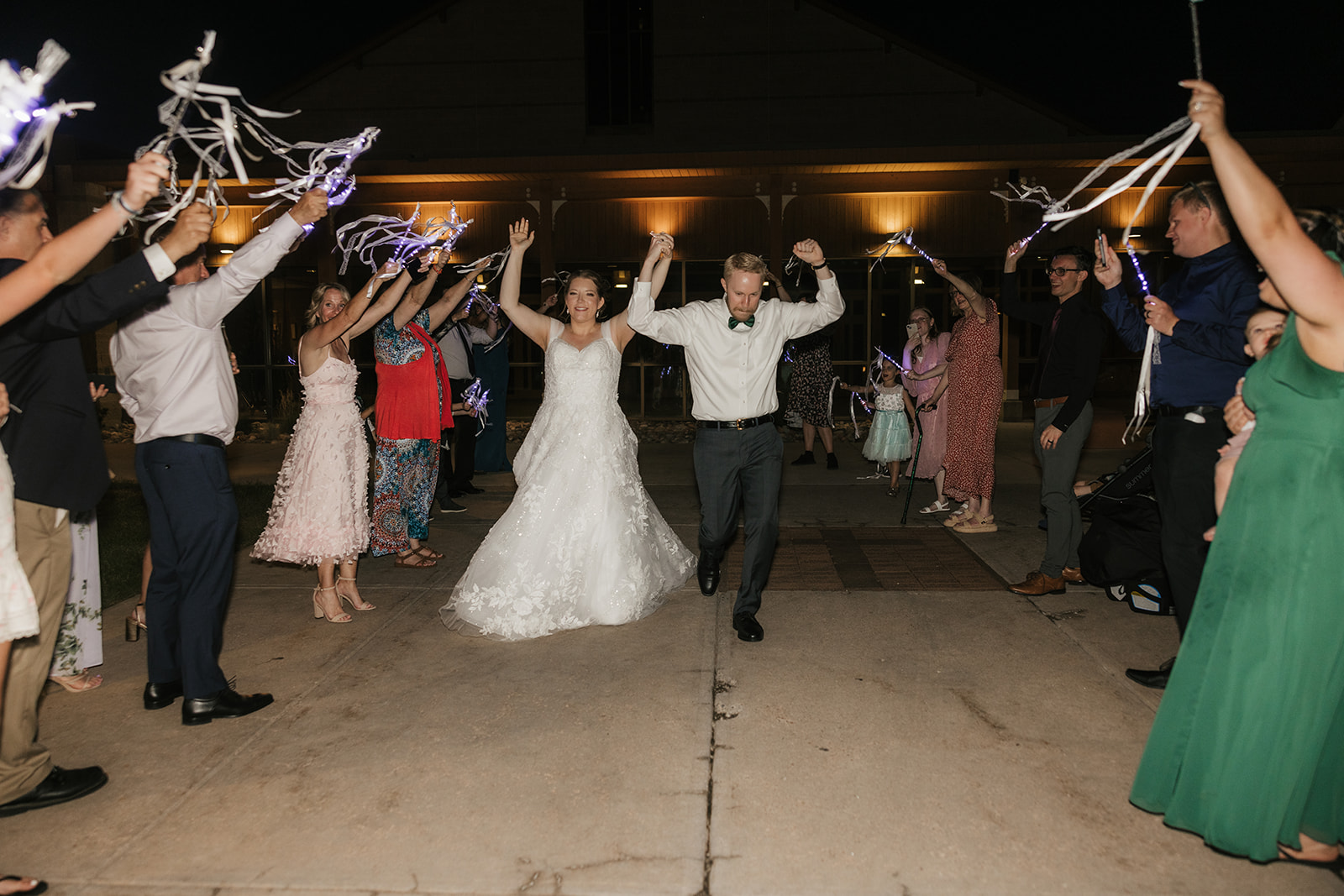 A bride and groom walk outside at night as guests hold up lights and ribbons, celebrating their wedding.
