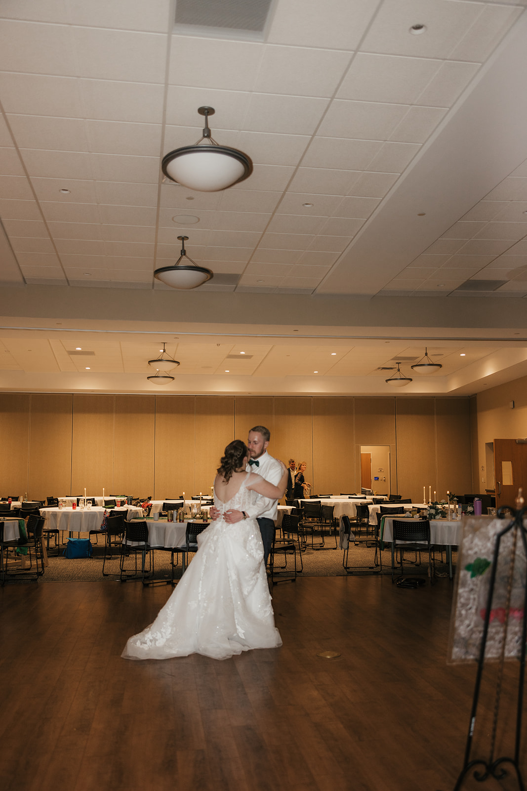 A bride and groom share a dance in the center of a large, decorated reception hall with tables and chairs arranged around them.