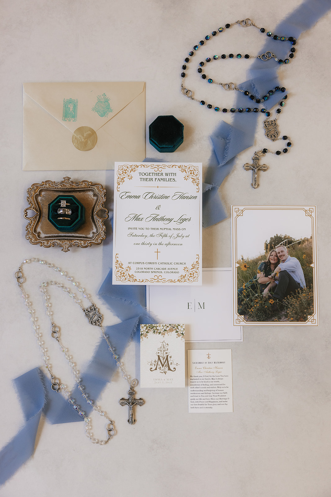A flat lay of wedding invitations, envelopes, a photo of a couple, a small framed picture, a ring box, and printed wedding materials arranged on a light surface with a ribbon.
