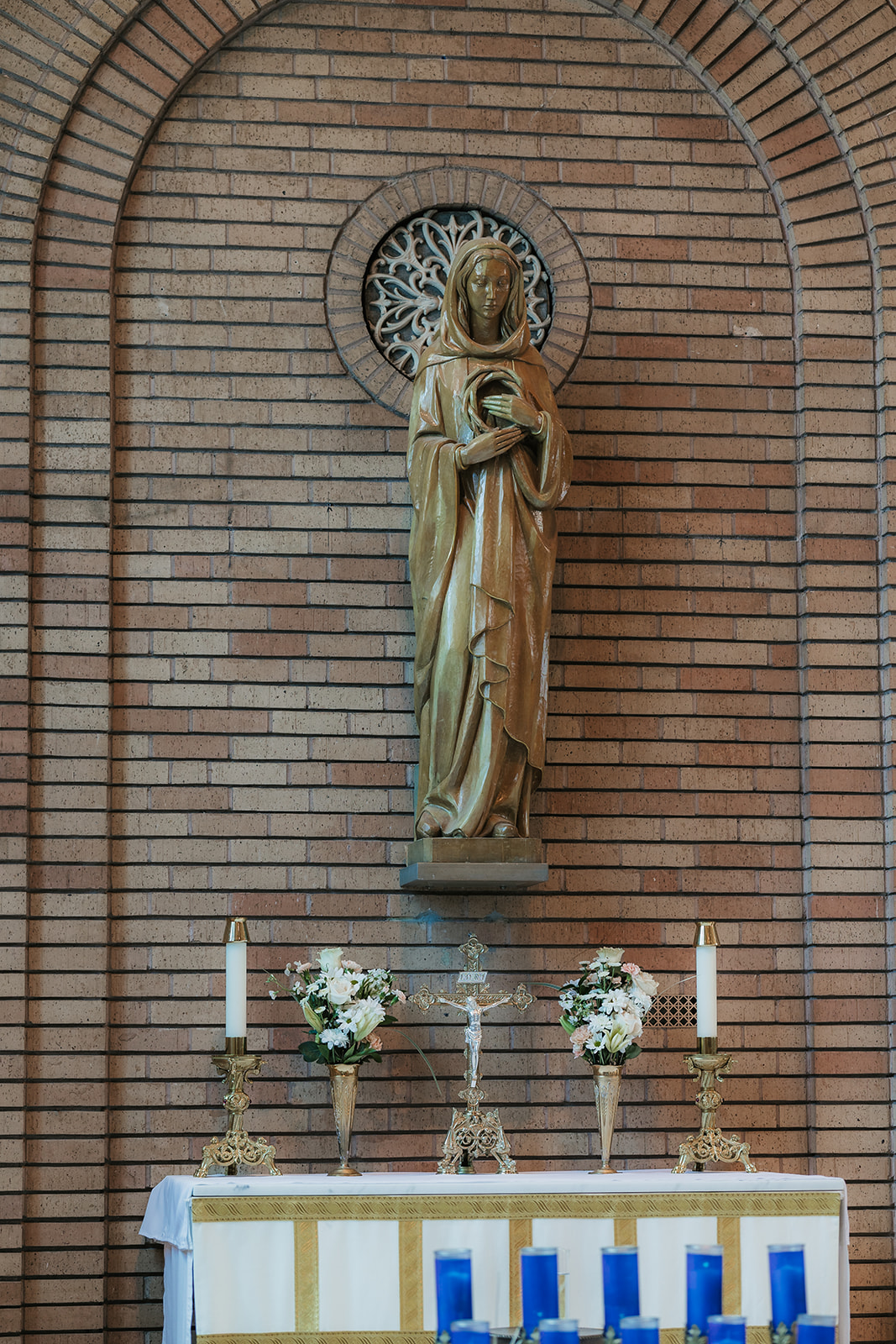 A statue of a robed figure stands in a brick alcove above an altar with flowers, candles, and a crucifix.