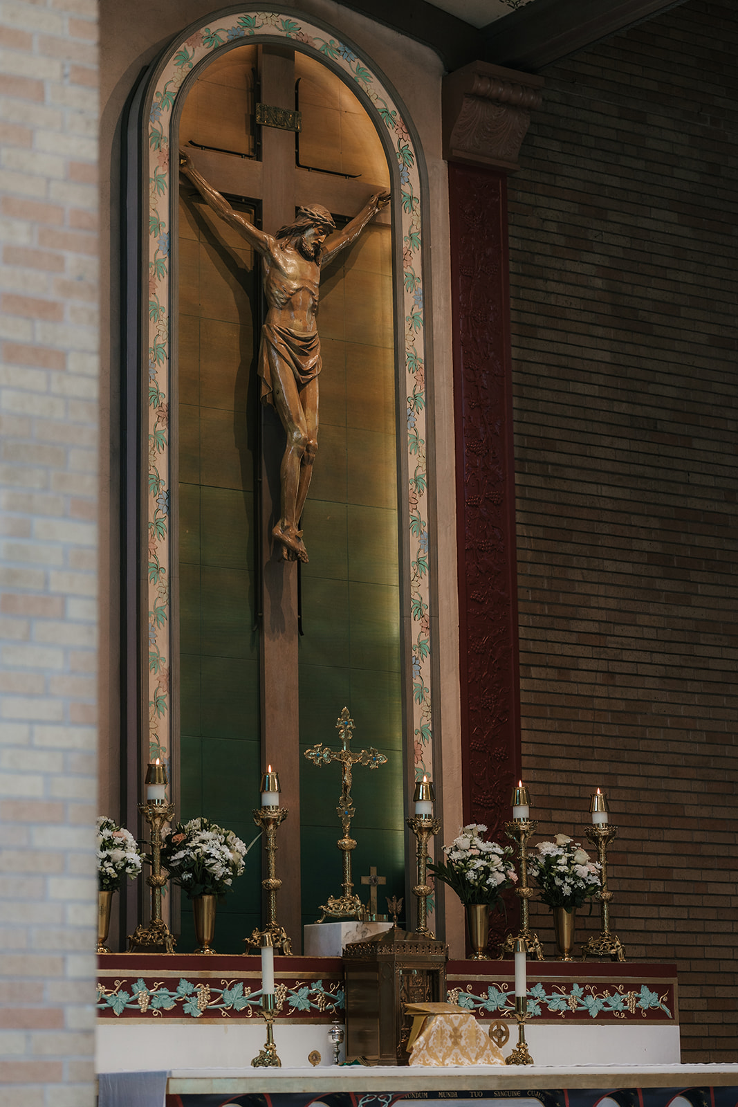 A statue of a robed figure stands in a brick alcove above an altar with flowers, candles, and a crucifix.