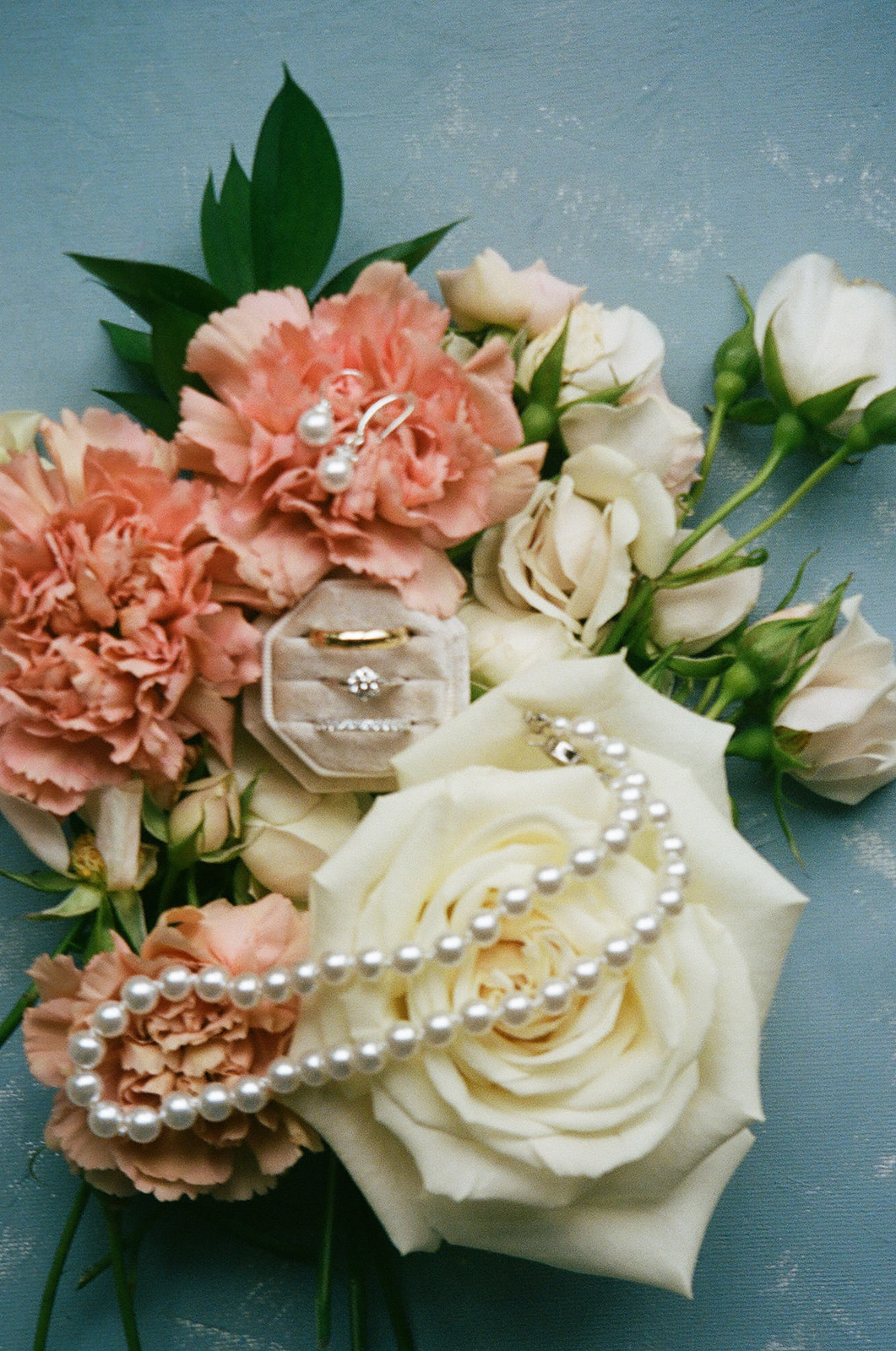 A pearl necklace, gold ring, and diamond earring rest on a large white rose, surrounded by pink and white flowers on a blue surface.