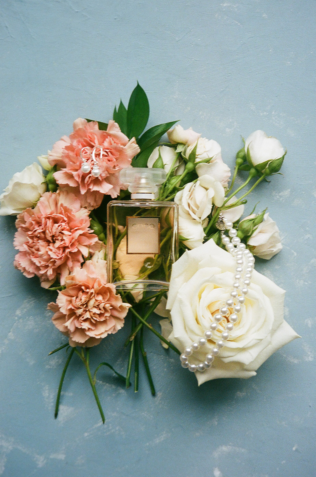 A pearl necklace, gold ring, and diamond earring rest on a large white rose, surrounded by pink and white flowers on a blue surface.