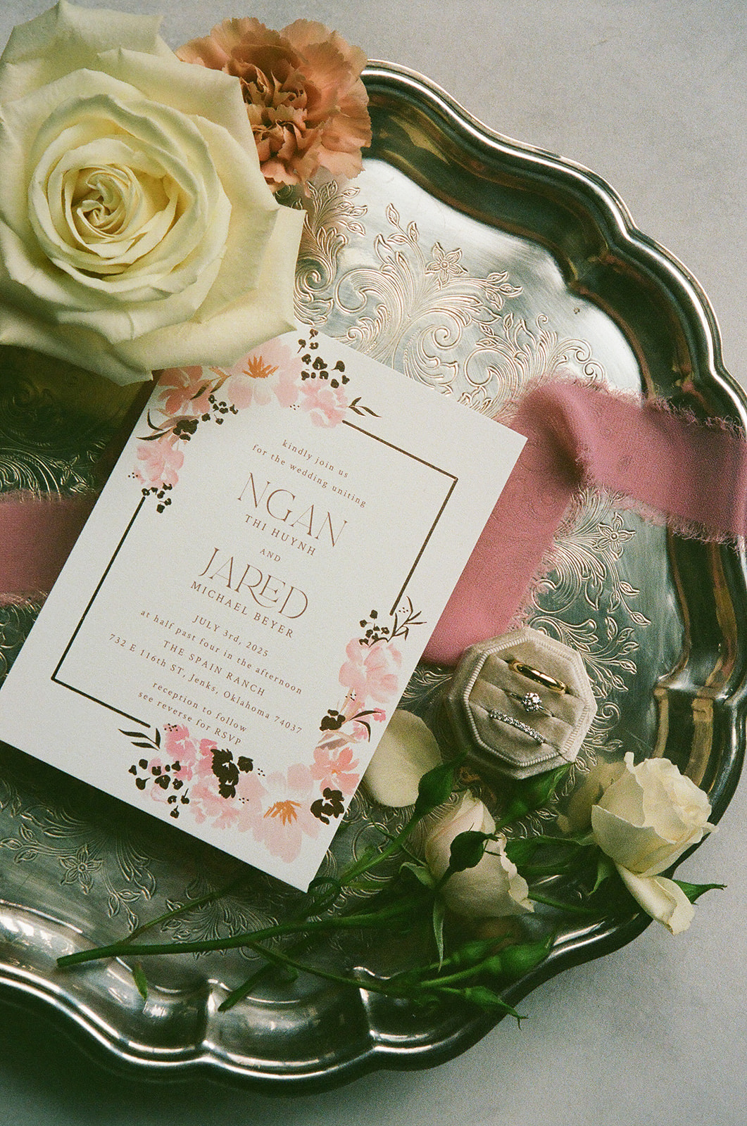 A pearl necklace, gold ring, and diamond earring rest on a large white rose, surrounded by pink and white flowers on a blue surface.