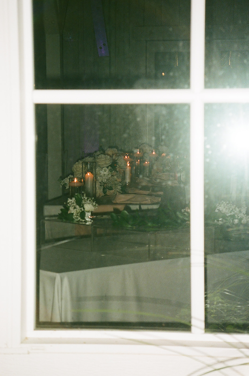 A long dining table set with plates, candles, and floral arrangements is positioned in a softly lit room with white walls; a tiered cake sits by the window in the background.