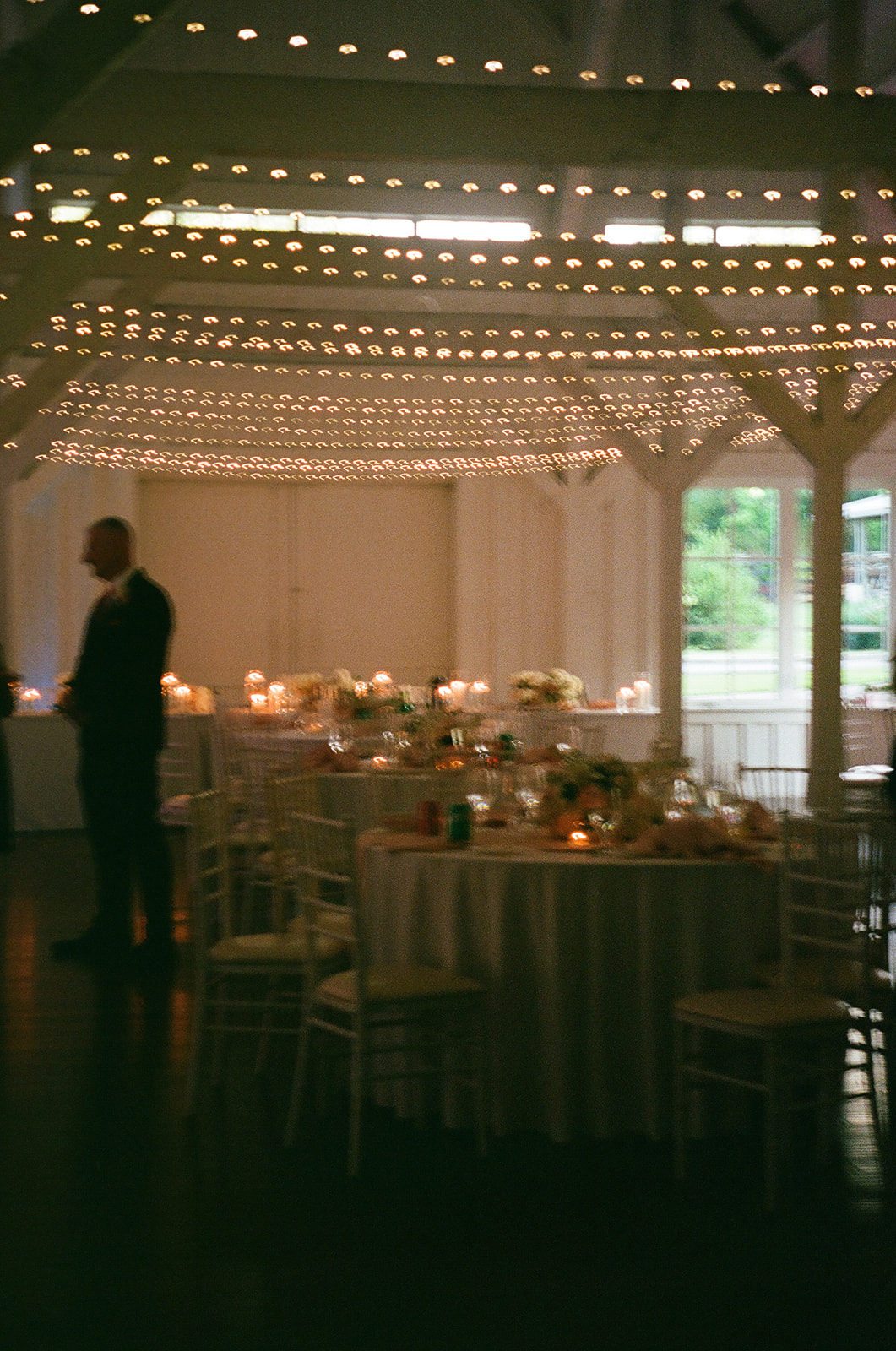 A long dining table set with plates, candles, and floral arrangements is positioned in a softly lit room with white walls; a tiered cake sits by the window in the background.
