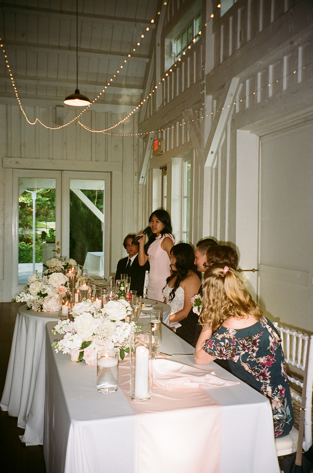 A long dining table set with plates, candles, and floral arrangements is positioned in a softly lit room with white walls; a tiered cake sits by the window in the background.