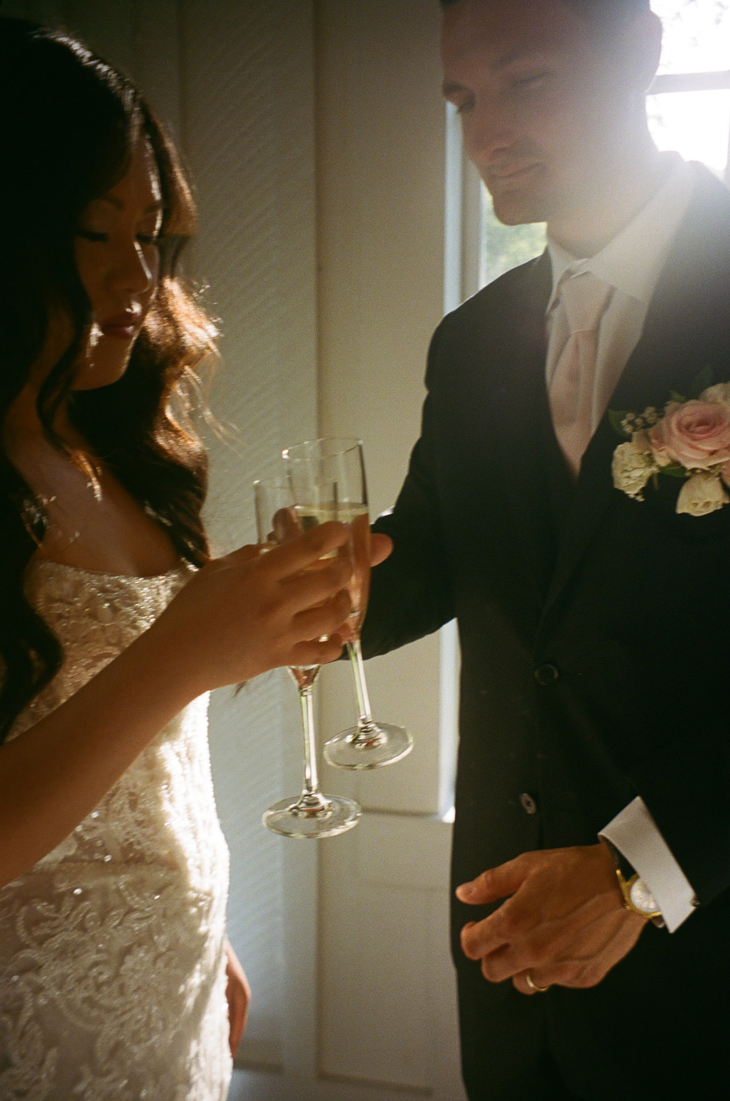 A bride and groom in formal attire clink champagne glasses, standing close together in soft natural light.