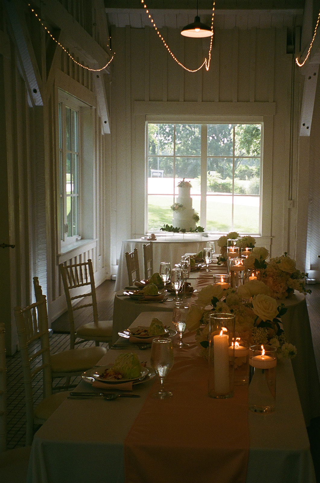 A long dining table set with plates, candles, and floral arrangements is positioned in a softly lit room with white walls; a tiered cake sits by the window in the background.