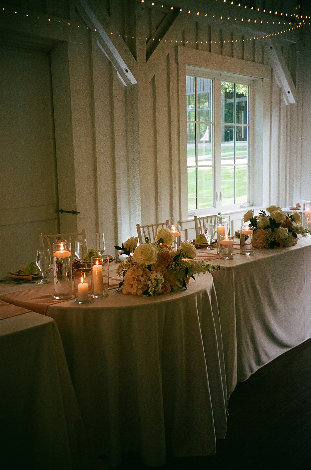 A long dining table set with plates, candles, and floral arrangements is positioned in a softly lit room with white walls; a tiered cake sits by the window in the background.