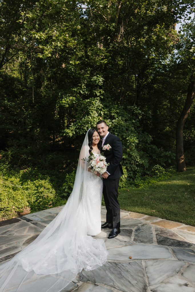 A bride and groom stand together outdoors on a stone path, surrounded by greenery. The bride holds a bouquet and wears a long white dress with a veil for a wedding at Spain Ranch