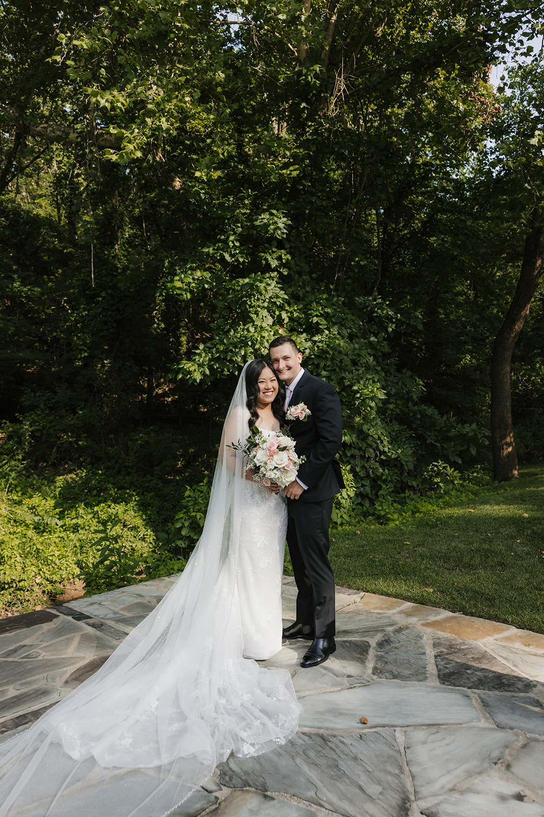 A bride and groom stand together outdoors on a stone path, surrounded by greenery. The bride holds a bouquet and wears a long white dress with a veil for a wedding at Spain Ranch