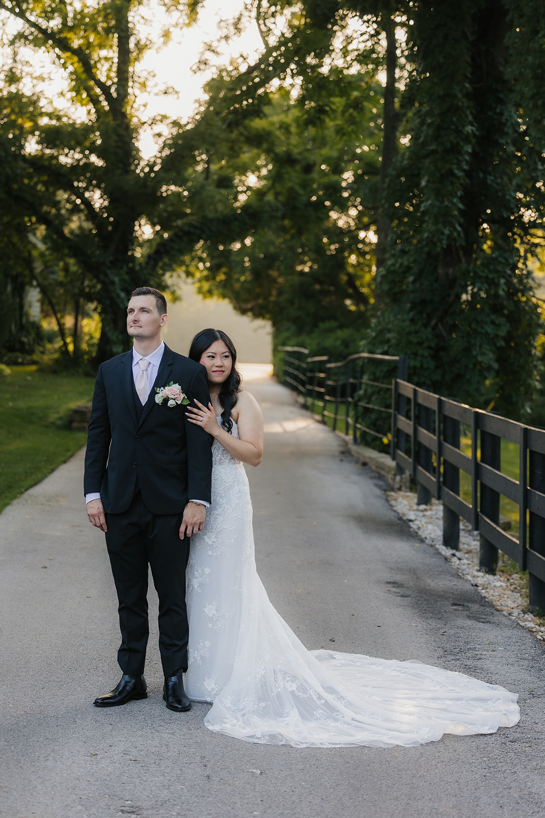 A bride and groom stand together outdoors on a stone path, surrounded by greenery. The bride holds a bouquet and wears a long white dress with a veil for a wedding at Spain Ranch