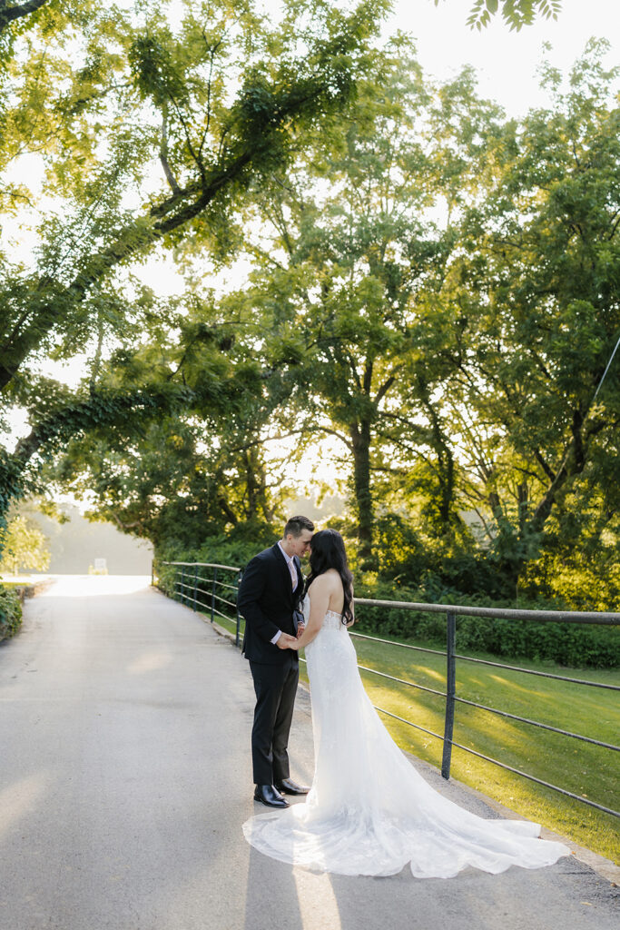 A bride and groom stand together outdoors on a stone path, surrounded by greenery. The bride holds a bouquet and wears a long white dress with a veil for a wedding at Spain Ranch
