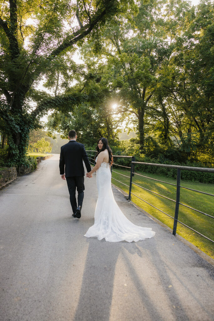 A bride and groom stand together outdoors on a stone path, surrounded by greenery. The bride holds a bouquet and wears a long white dress with a veil for a wedding at Spain Ranch