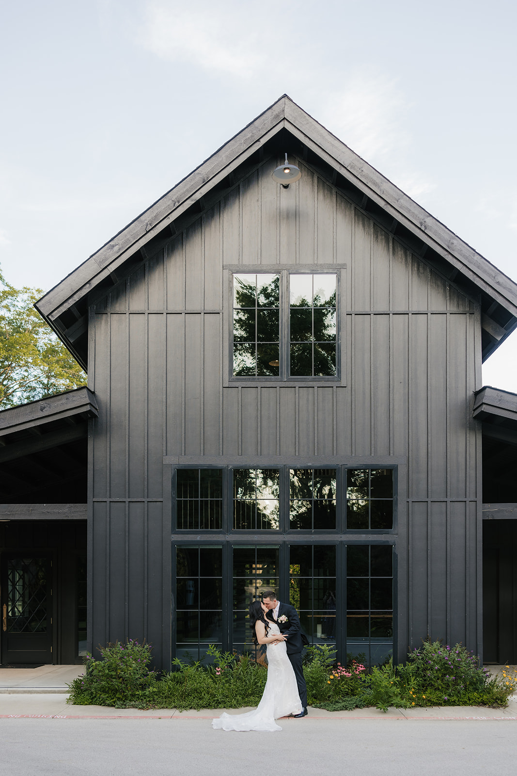 A bride and groom stand embracing in front of a modern black barn-style building with large windows and gable roof.