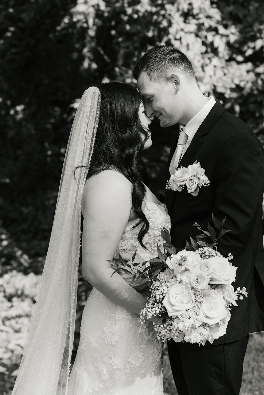 A bride and groom stand together outdoors on a stone path, surrounded by greenery. The bride holds a bouquet and wears a long white dress with a veil for a wedding at Spain Ranch