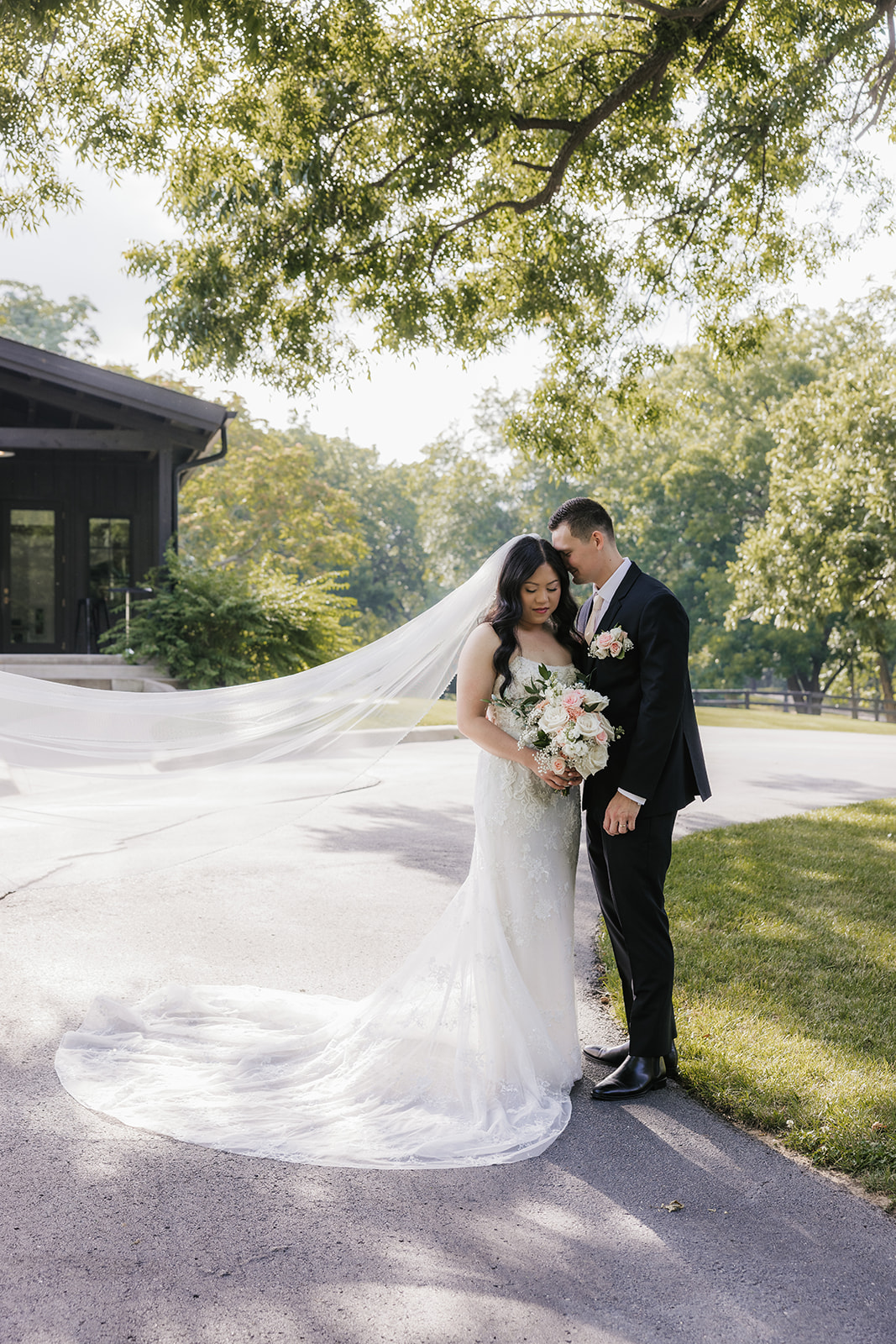A bride and groom stand together outdoors on a stone path, surrounded by greenery. The bride holds a bouquet and wears a long white dress with a veil for a wedding at Spain Ranch