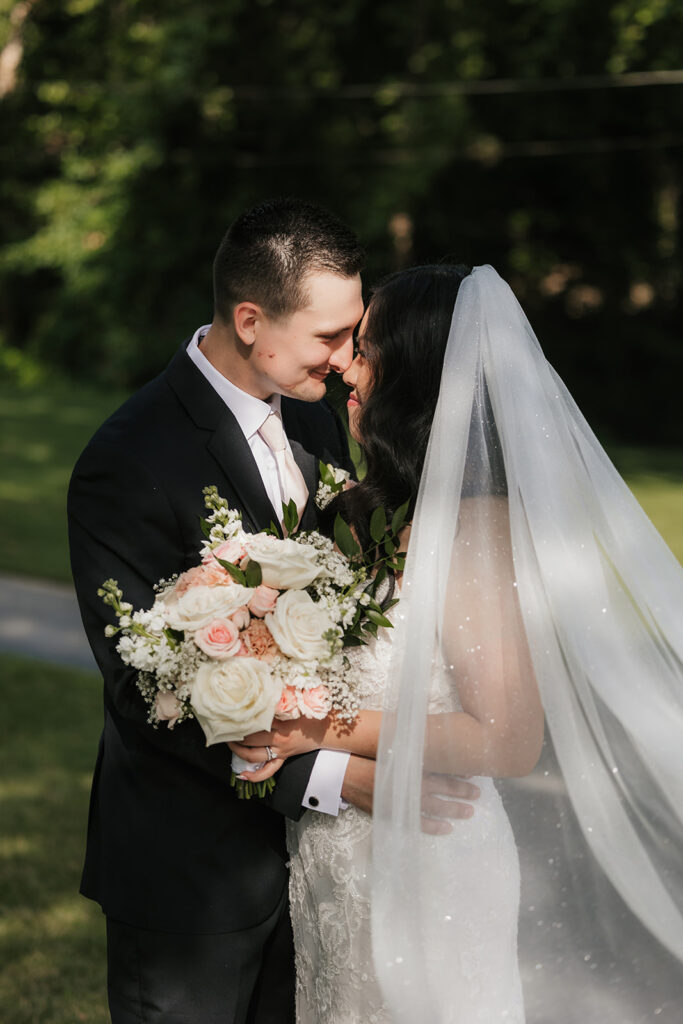 A bride and groom stand together outdoors on a stone path, surrounded by greenery. The bride holds a bouquet and wears a long white dress with a veil for a wedding at Spain Ranch