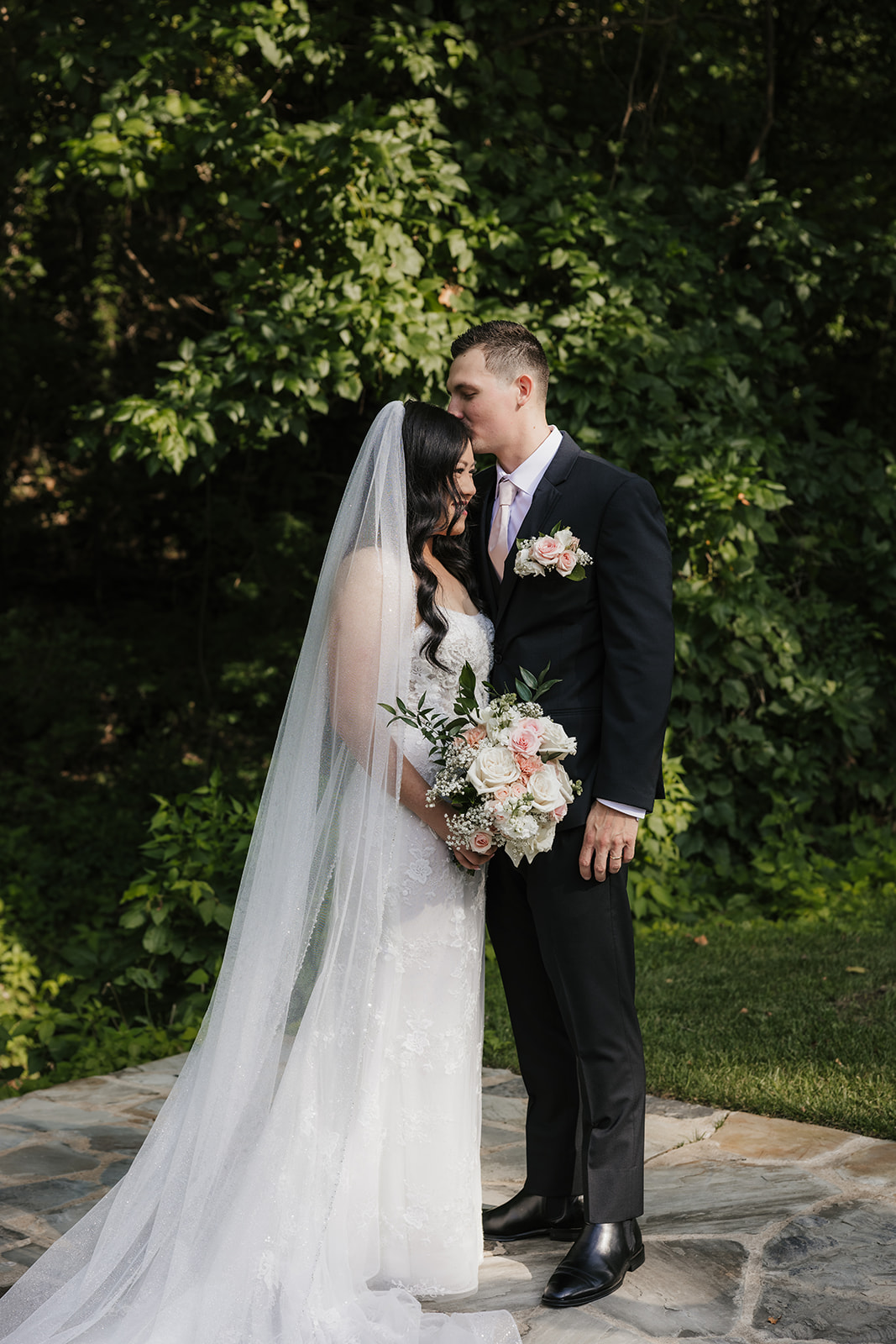 A bride and groom stand together outdoors on a stone path, surrounded by greenery. The bride holds a bouquet and wears a long white dress with a veil for a wedding at Spain Ranch
