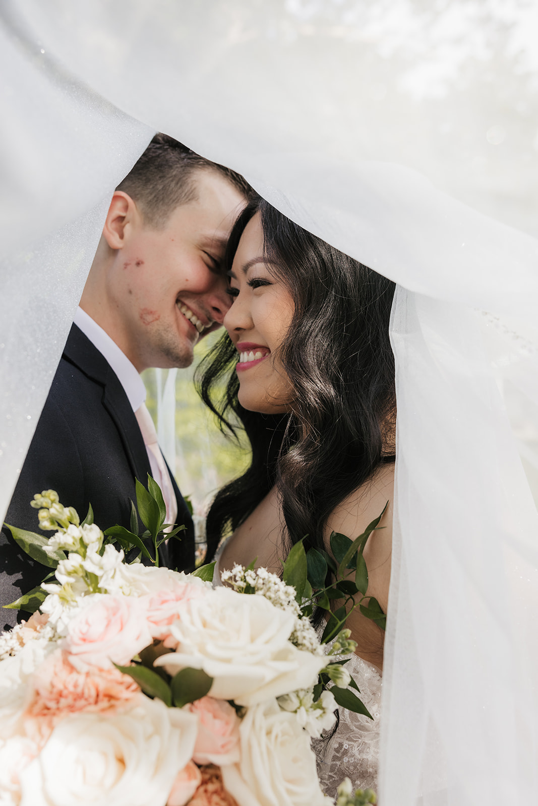 A bride and groom stand together outdoors on a stone path, surrounded by greenery. The bride holds a bouquet and wears a long white dress with a veil for a wedding at Spain Ranch