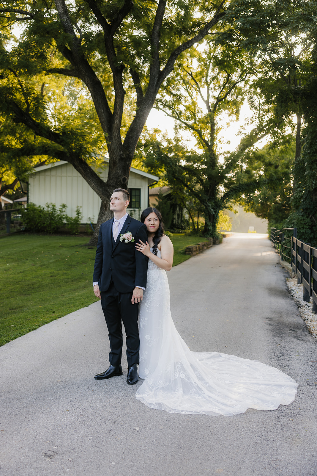 A bride and groom stand together outdoors on a stone path, surrounded by greenery. The bride holds a bouquet and wears a long white dress with a veil for a wedding at Spain Ranch