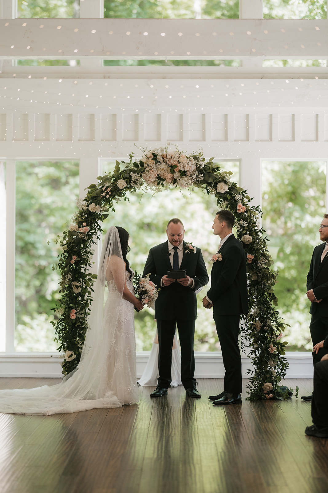 A bride and groom stand with an officiant under a floral arch indoors, exchanging vows during a wedding ceremony at spain ranch