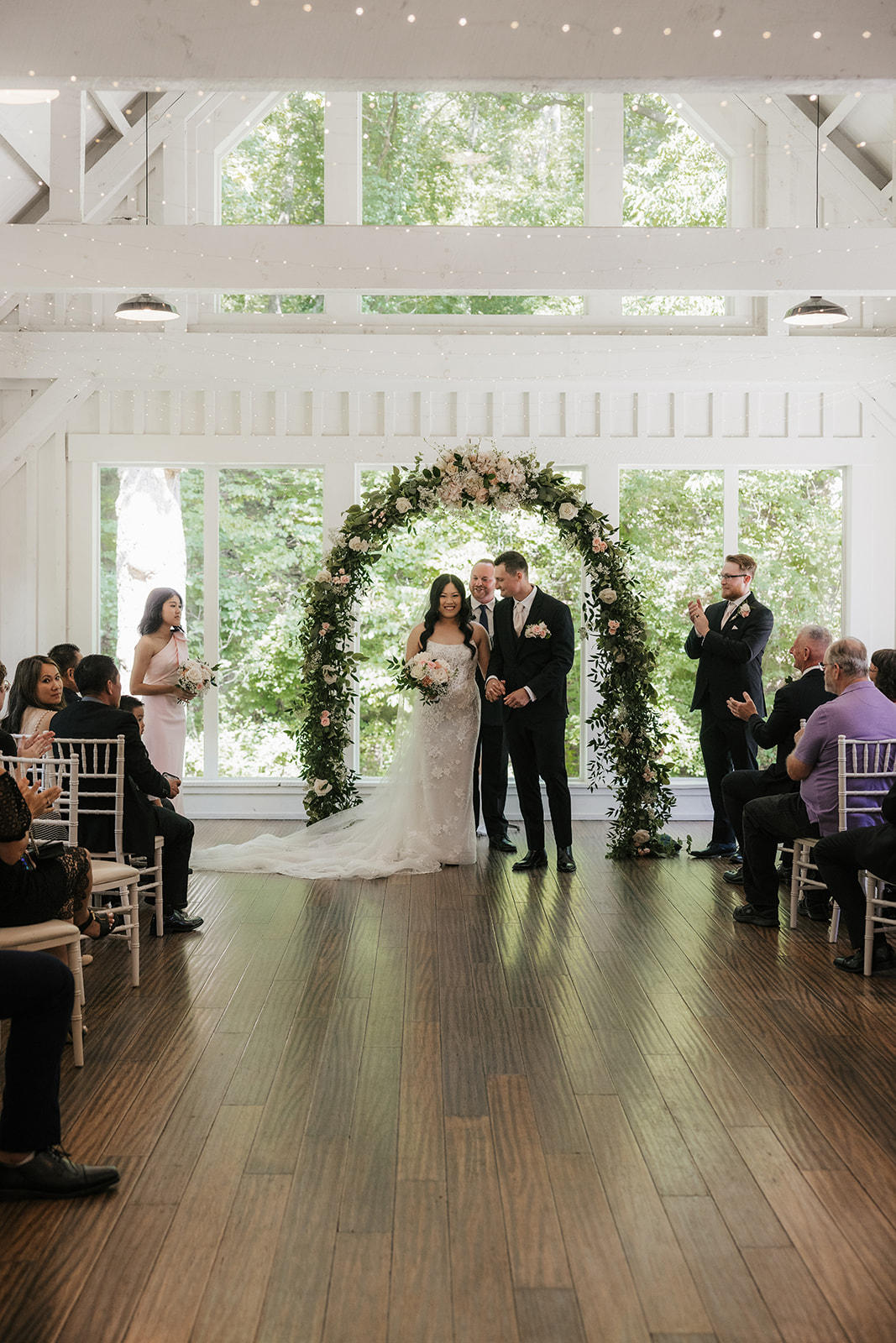 A bride and groom stand with an officiant under a floral arch indoors, exchanging vows during a wedding ceremony at spain ranch