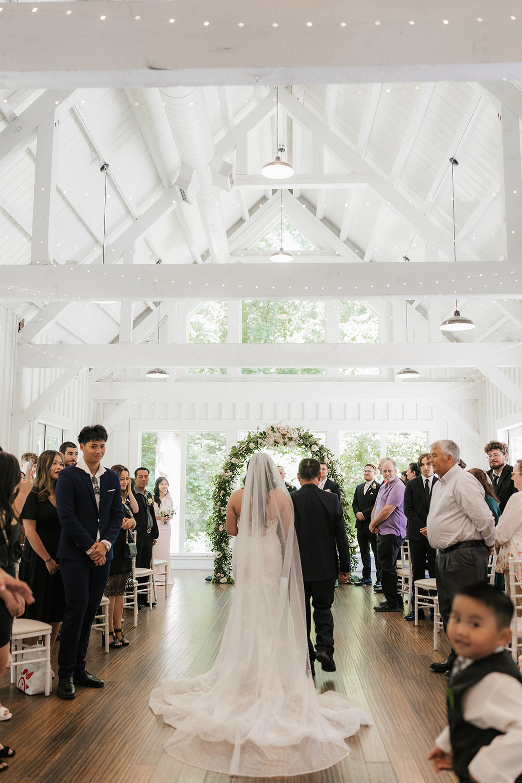 A bride and groom stand with an officiant under a floral arch indoors, exchanging vows during a wedding ceremony at spain ranch