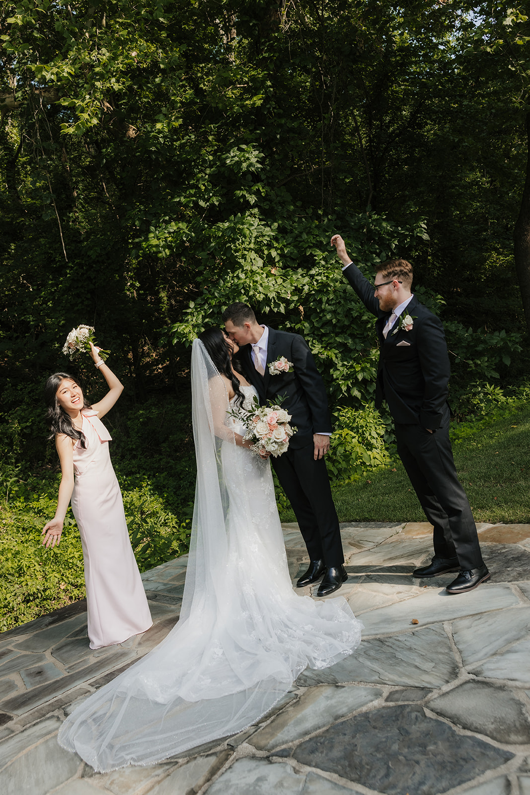 A bride and groom kiss outdoors on stone pavement while a bridesmaid and groomsman cheer beside them. Trees fill the background.
