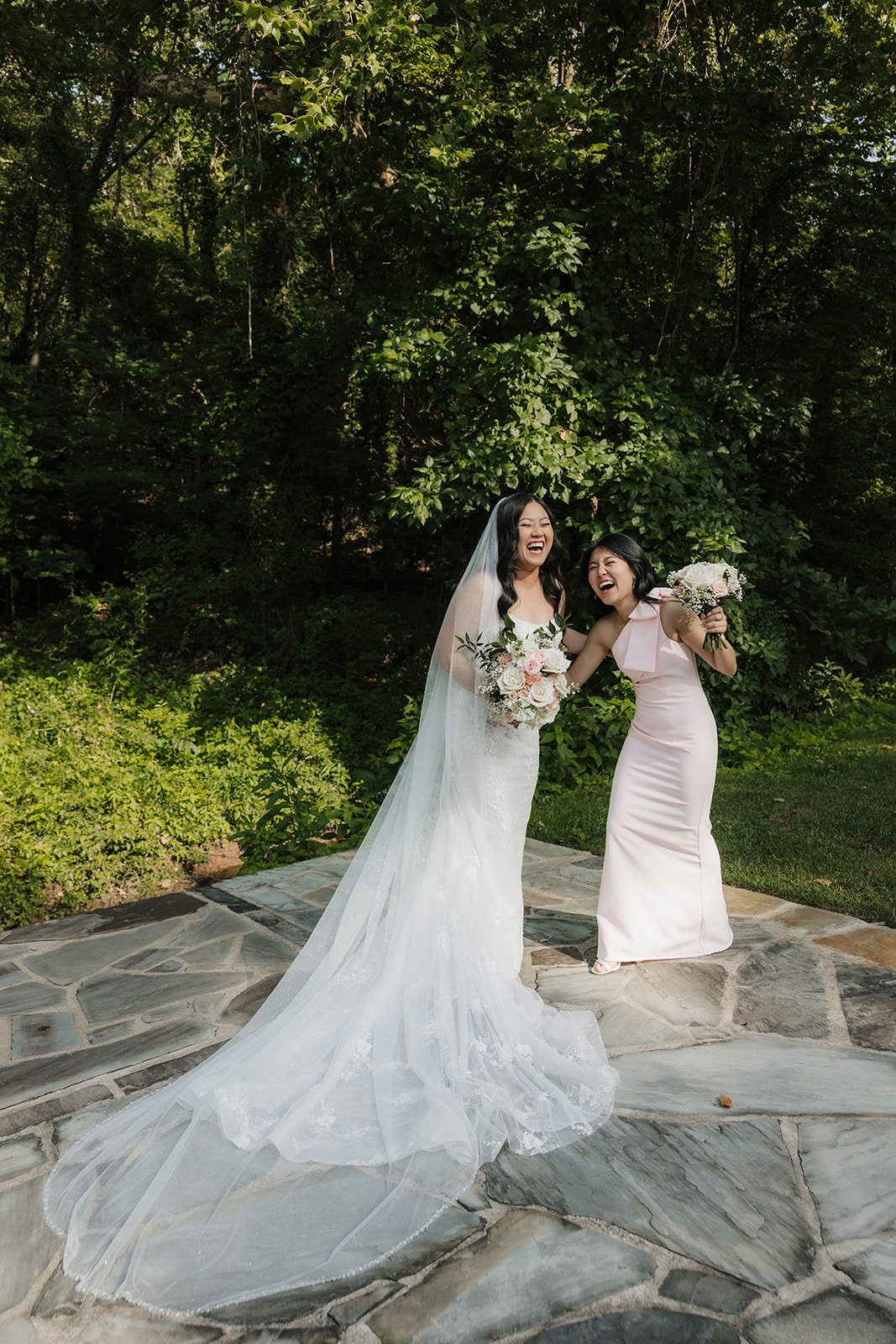A bride and groom kiss outdoors on stone pavement while a bridesmaid and groomsman cheer beside them. Trees fill the background.
