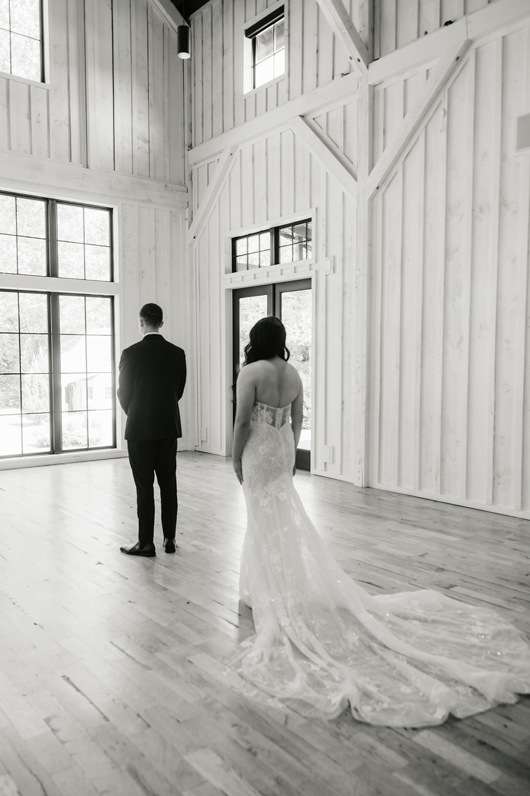 A bride in a wedding dress approaches a groom from behind inside a bright, spacious venue with high ceilings and large windows.