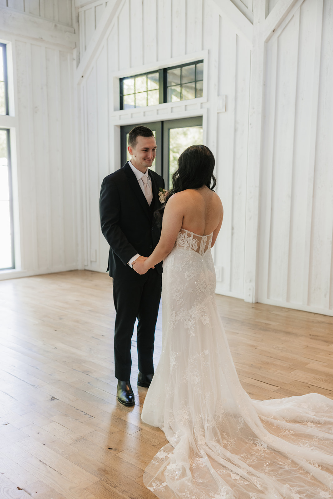 A bride in a wedding dress approaches a groom from behind inside a bright, spacious venue with high ceilings and large windows.